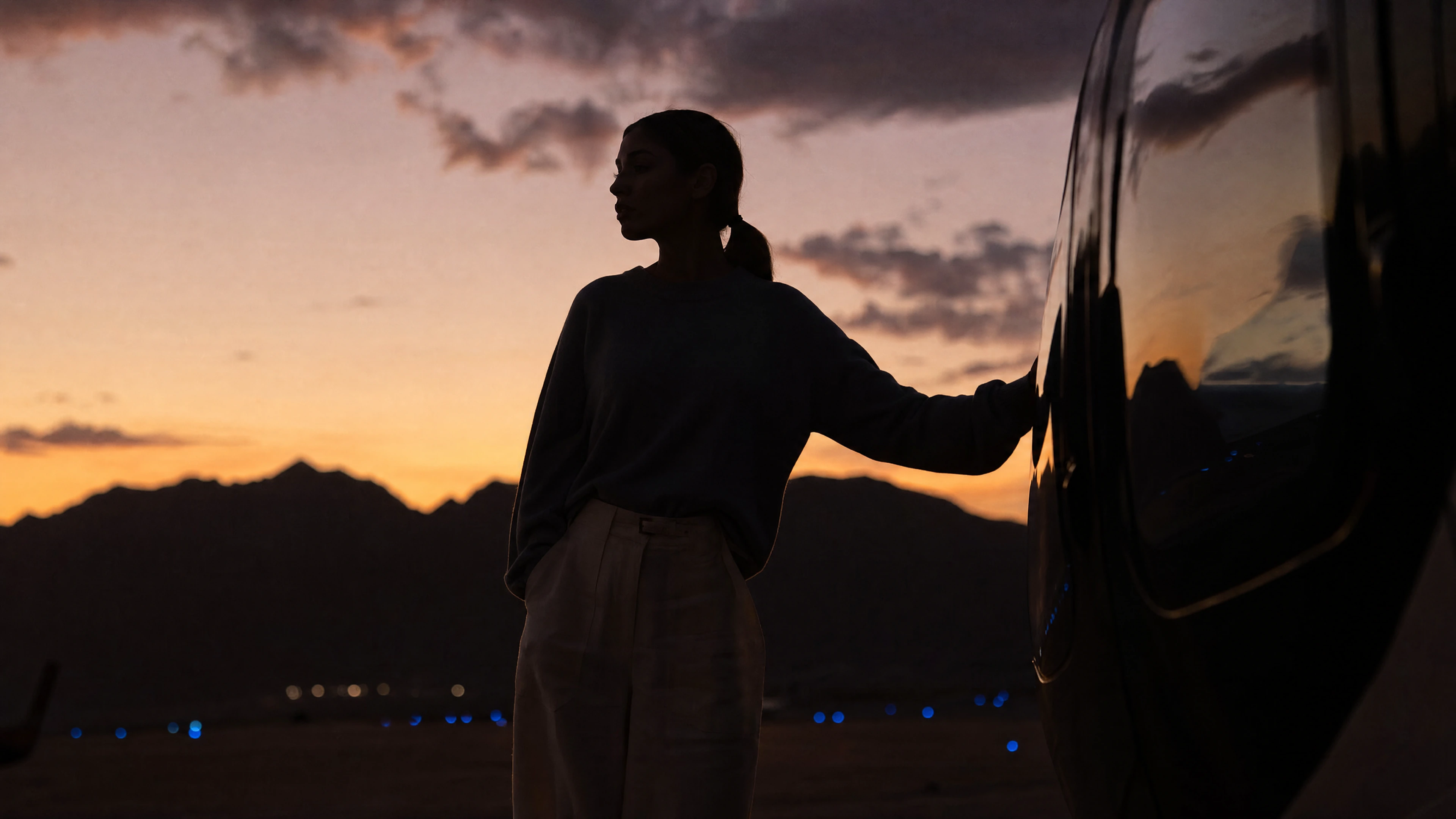 Woman resting her hand on a dark SUV at dusk in the desert, with mountains and a sunset sky behind her